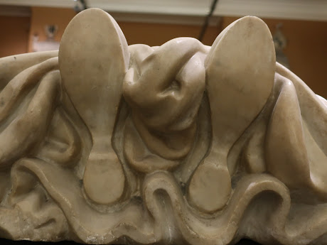 Photo. A stone effigy that is placed on a plinth in a museum, view of the effigy's feet and shoes, carved white stone, including draperies or hemline of a woman's dress.