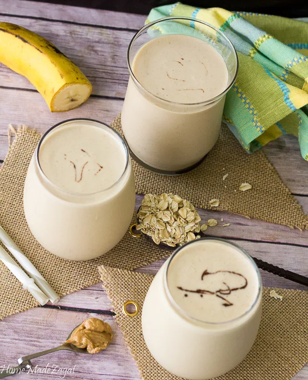 This picture is 3 glasses of peanut punch with a banana and some oats on the side.