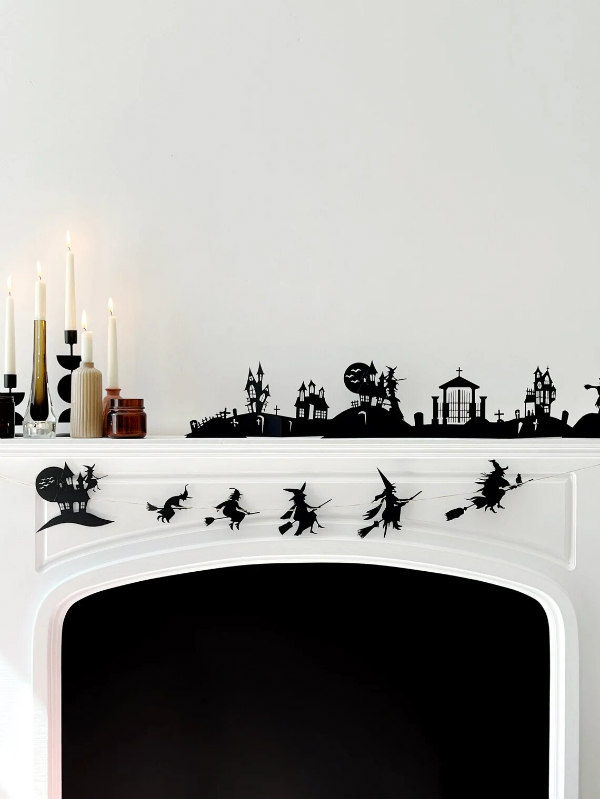 fireplace with mantel on which black silhouette paper cuttings of Halloween scene and witch garland are displayed