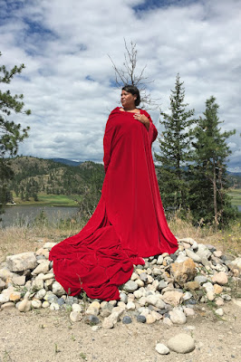 Indigenous woman wearing red flowing dress, standing on rocks, with a lake and trees in background