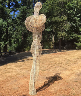 A tall thin humanoid figure of open basketweave stands very straight on brown grass.