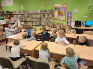 Pani bibliotekarka siedzi naprzeciwko dzieci i pokazuje im książkę. Tło: sala biblioteczna.