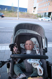 Kuva ulkoa bussipysäkin katoksesta kuvan ottaja tietä kohti. Tien toisella puolella näkyy suuri rakennus. Ilse istuu harmaissa rattaissa, kasvot poispäin tiestä. Katse suuntautuu ylöspäin (kuvan ottajan siis voi olettaa luultavasti istuvan).