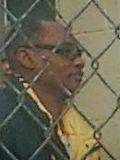 Snapshot taken through a chain link fence of a balding middle-aged Black man wearing eyeglasses.