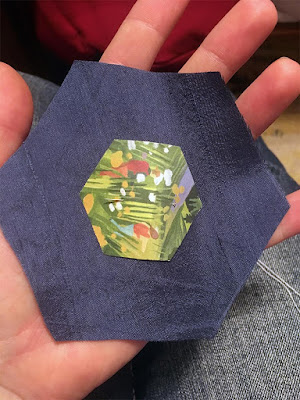 A close-up of a paper hexagon with a rather impressionist painting of a lush summer meadow, centered on a medium-blue silk hexagon, on a white hand with fingers spread to support the fabric.