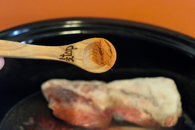 A slow measuring spoon with cayenne pepper, over the slow cooker with the pork in it.