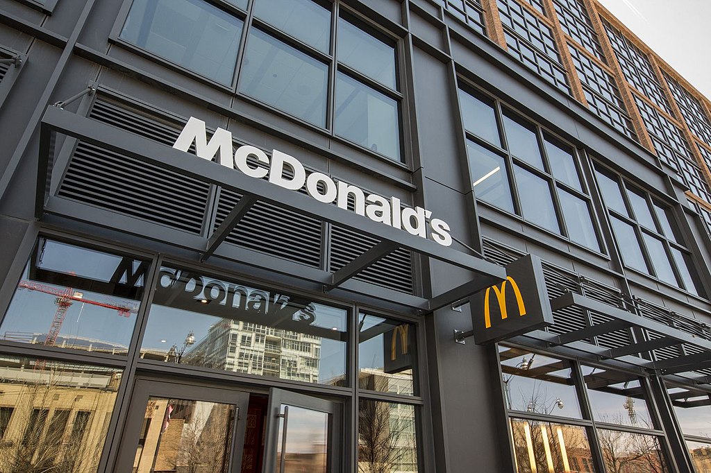 A Fast-Food chain McDonald’s is in an urban city environment, during the lunch rush. With an impressive open sky-line overlooking the city.