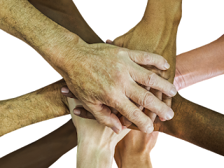 9 hands of various skin tones, clasped one atop the other, viewed from above, with some forearm visible for each.