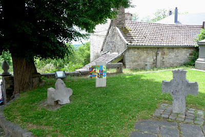 Stèle en béton plantée verticalement dans le sol et peinte avec des fromes colorées aux teintes crayeuses. Un volet en bois est accroché sur la gauche peint avec des formes plus petites.