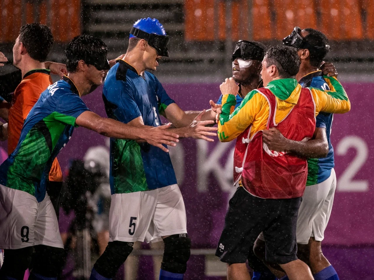 Os jogadores brasileiros, de camisa azul e calções brancos, se reúnem debaixo de chuva para comemorar o gol. Na imagem, Cássio e Tiago, dois homens brancos, estão de braços abertos para abraçar Jeffinho, homem preto, que vai até ao banco de reservas para comemorar com os colegas