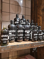 A wide shot photo of a bunch of cylindrical bottles filled with green liquid with a black label with Grass Shower Gel lush in white fonton a bright background