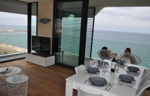 salon con mesa blanca y cuatro sillas junto a una televisión y grandes ventanales con vistas a la playa de mataro 