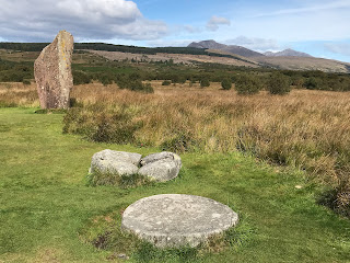 A photo of some circular grey and flat stones on Machrie Moor with a red sandstone standing stone behind them.   Photograph by Kevin Nosferatu for the Skulferatu Project.