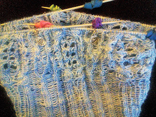 A knit hat with live stitches on double-pointed needles, knit in a cable and lace pattern.   The needles have elastics on the end.  The yarn is a pale blue fingering-weight yarn.