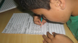 Imagem de um menino baixa-visão, cabeça encostada próximo a carteira, escrevendo numa folha de pautas ampliadas