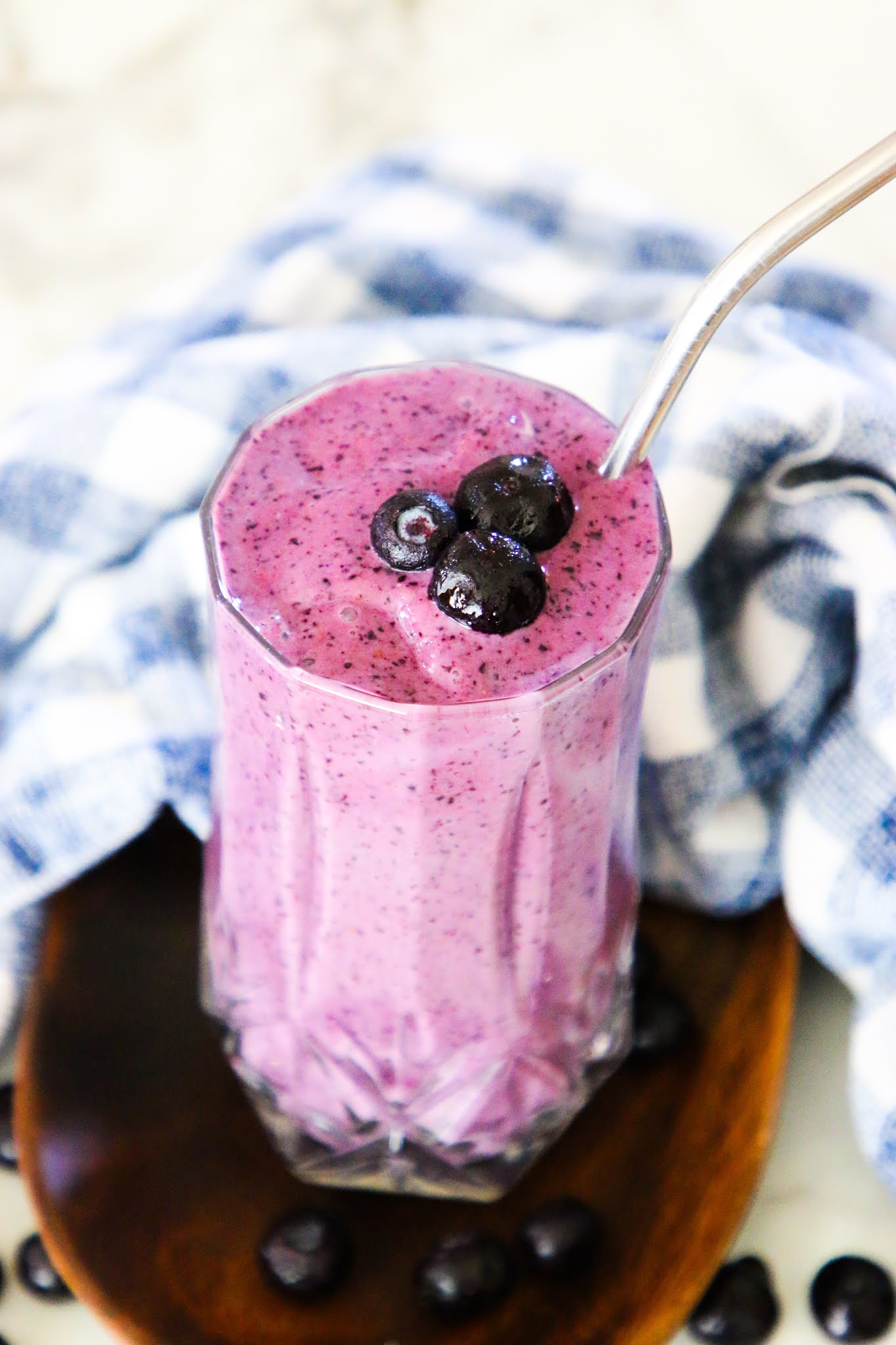 Purple blueberry smoothie in a tall glass with blueberries on top. There is a wood coaster underneath the smoothie sitting on top of a white marble table. The glass is wrapped in a blue dishcloth.