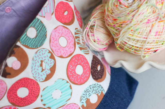 Donut print and denim boxed base project bag with a lilac interior and light pink snap buttons on the top. It's next to a cake plate with a speckled cake of yarn and speckled hank of yarn.