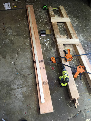 A high-angle shot of a long board with a much smaller strip screwed to it along the length, a few inches from the right edge, laid parallel to two 4x4s with small 2x4 offcuts resting across them. A cordless drill, two orange-and-black clamps, a box of screws, a green tape measure, and a small stack of envelopes with a black pen and yellow pencil are scattered around the sawdusty grey concrete floor.