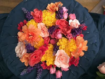 A photo from above of an upturned straw hat, lined with medium-blue pleated fabric held in place with silver pins, and with the crown completely filled and overflowing with bright-yellow, peach, pale-pink, hot-pink, and putple fake flowers. There are chrysanthemums, two rose types, anemones, lavender, and oriental poppies.