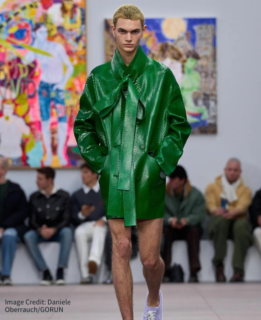 Photo of model in Loewe runway. Model is wearing forest green textured jacket with bow tied around the neck, no trousers and a pair of lilac tennis shoes