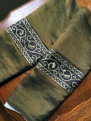 A pair of green-gold sleeves, divided in the center with bands of gold floral embroidery on brown silk, laid flat and slightly overlapping on a wooden table.