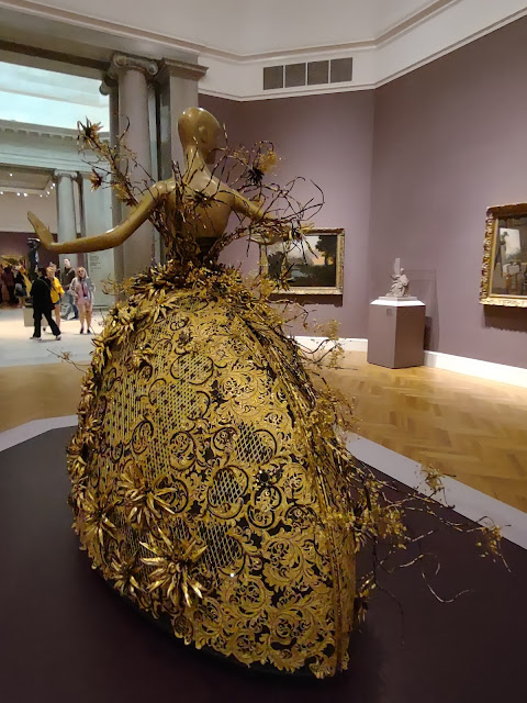 Photo of a spectacular gold dress on a slender mannequin. The dress is seen from the rear. It is architectural, a semi-circle flaring from front to back and slightly to the sides. It looks like a tapestry, with black underneath and gold on top, with big appliqued sunbursts and other shapes. Black and gold branchlike decorations arise from the mannequin's waist.