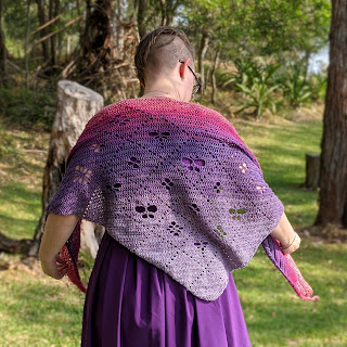 Sarah is standing with back to the camera looking down, her arms are raised slightly as she rearranges her shawl. It is crochet cotton yarn in a gradient from pale purple to dark purple to pink. It has filet design of a grid with motifs of flowers, butterflies and dragonflies. She is standing in a grassy clearing surrounded by trees.