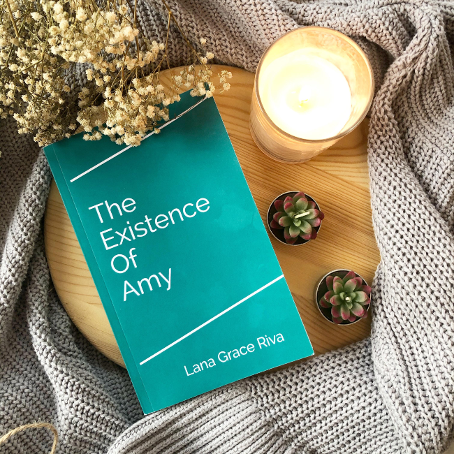 A plain teal book cover for 'The Existence of Amy' on a brown chopping board on top of a grey cardigan, with baby's breath flowers