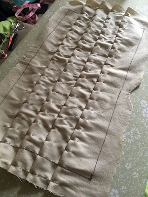 A beige rectangle of fabric with the edges marked with black sharpie and large seam allowances left outside the marks, with four straight pleats pinned across the edges about every half inch. The pleats are neat at the edges, but heavily puckered between the pins. The pile of sewing supplies on the couch, just visible, has grown to large silver shears, their black plastic sheath, a spool of red thread, and an uncoiled pink measuring tape.