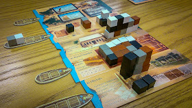 The five main play boards, each with a variety of blocks, in white, black, brown, and grey, stacked on them in different ways. The Market Board is the exception; it has no blocks, but does have some market cards on it. The boat tokens are docked with four of the five boards by having their prow placed in the notch on the left side of the board. All the blocks that were on these boats have been moved to the play boards, except a white one and a grey one on the second boat which haven't yet been transferred.