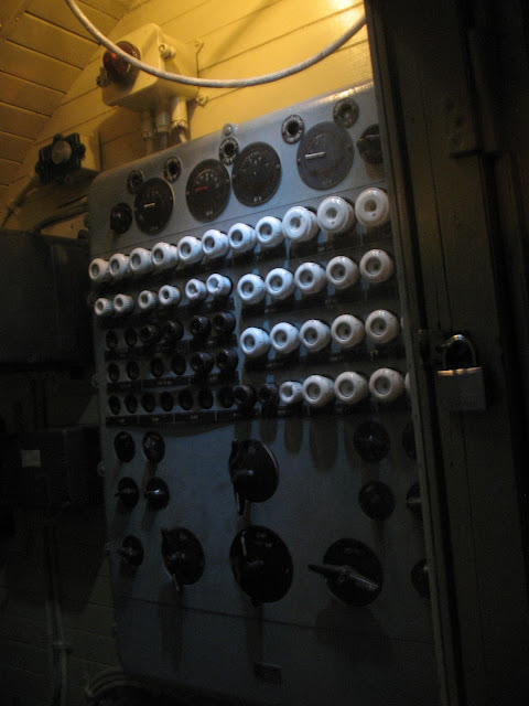 Photo of a WWII era submarine control panel. The panel is gray and has gauges at the top and bottom. In between are two rows of protruding white objects, below the white objects are protruding black objects.