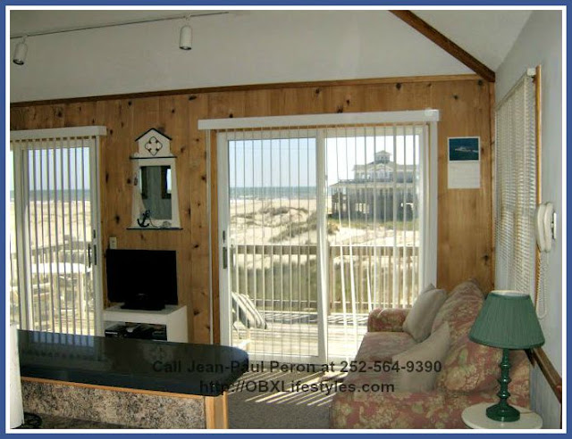 The fully carpeted living room of this 3 bedroom Outer Banks NC home for sale has large windows and sliding glass doors that allow natural light to stream in. 
