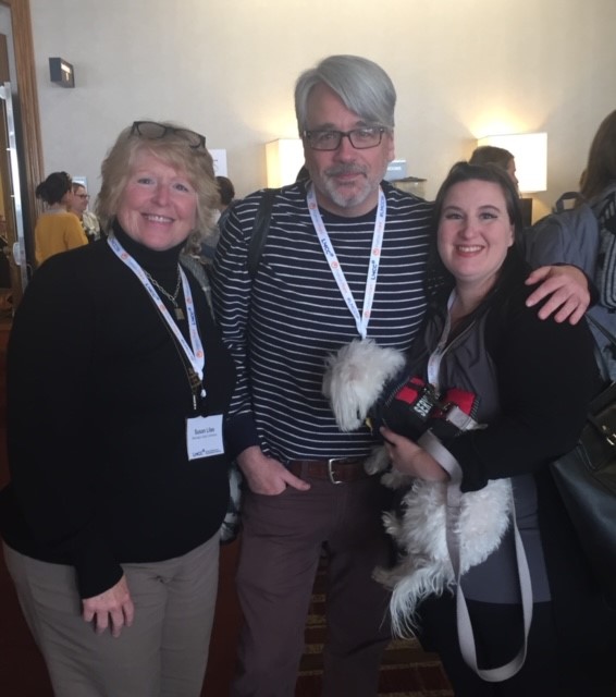 2 smiling women lean into smiling man in middle 1 woman is holding a service dog