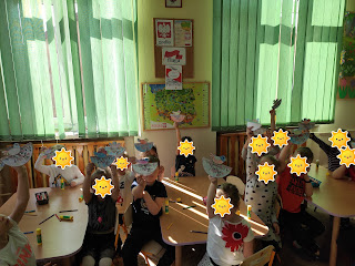 Na tle okna w sali przedszkolnej siedzą przy stoliku dzieci i prezentują wykonaną pracę plastyczną, tzn. trójwymiarowego ptaszka wykonanego z papieru i pokolorowanego kredkami przez dzieci