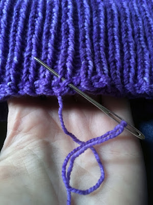 A close-up of the edge of a section of purple knitted ribbing, with a silver yarn needle threaded with the free end of yarn and inserted under two stitches.