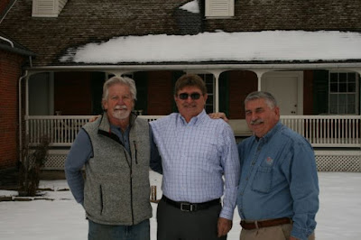 Three white men stand together with snow on the ground, there is snow on the roof of the rear porch of the house behind them. Man on left has gray hair and a gray goatee and mustache and is wearing a long-sleeved blue shirt under a gray zippered vest. Man in middle has brown hair and is wearing sunglasses and a white checked shirt. Man on right has graying hair and mustache and is wearing a blue denim shirt with "Old Economy Village" over the left pocket.