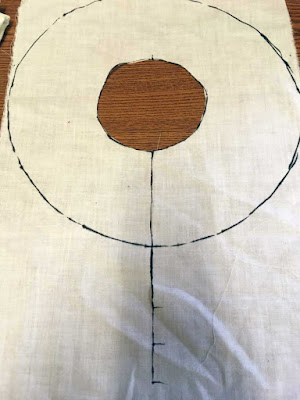 A rectangle of near-white fabric with torn edges, a small circle cut from the center, and a larger circle marked in black sharpie, with a line connecting the two circles' lowest points and continuing downward, with hashmarks at regular intervals on the descender.