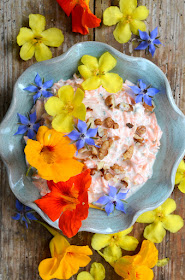 Cremiger Karottensalat mit Joghurt in einer grünblauen Schüssel mit gewelltem Rand. Rundherum und auf dem Salat liegen Blüten in den Farben gelb, orange und blau.