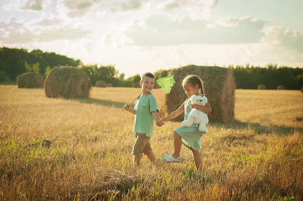 Dois amigos, uma menina e um nenino a brincar no  campo ao entardecer. #PraCegoVer