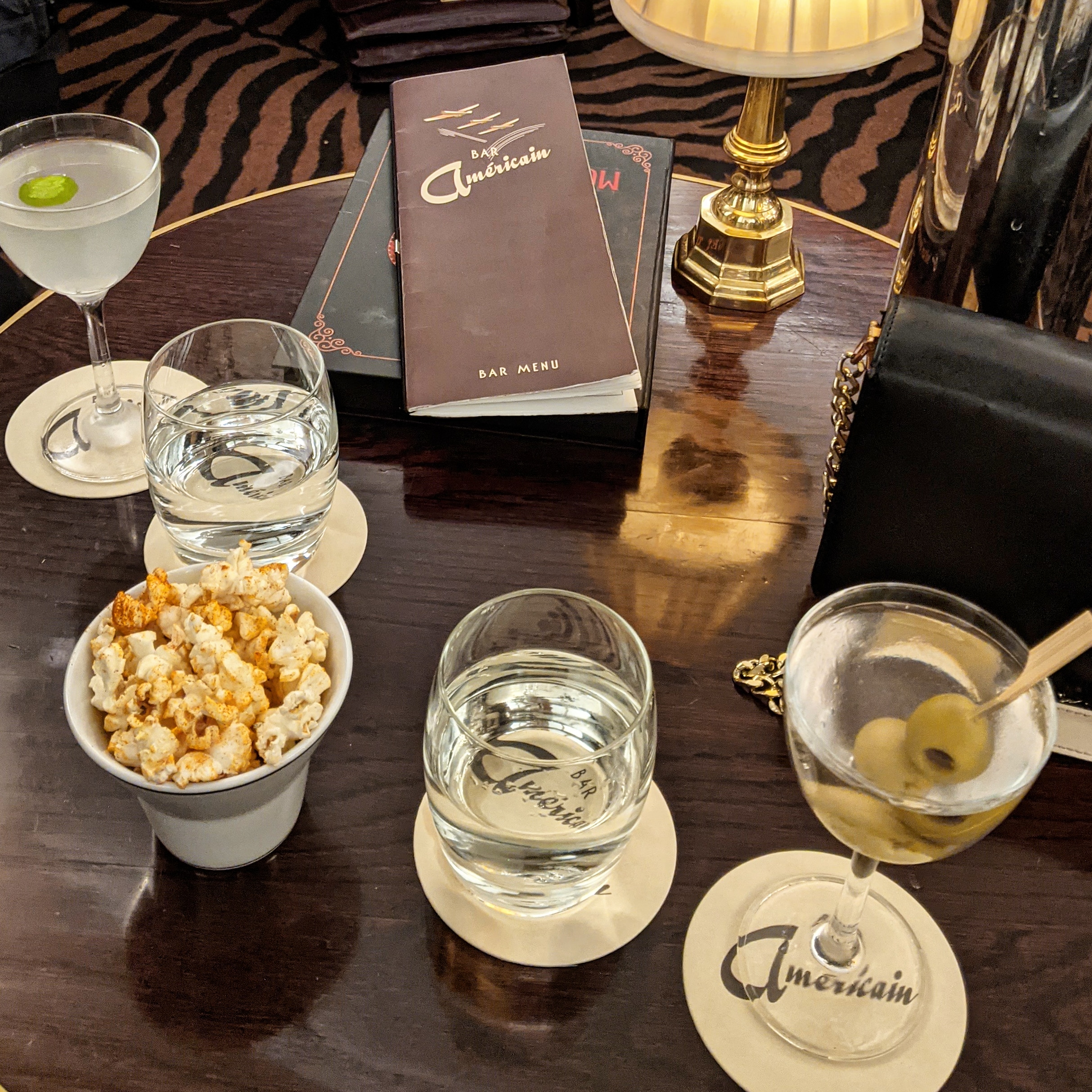 one of the best cocktails in london can be found at the stunning bar americain inside brasserie zedel, the image shows a wooden table topped with two cocktails and a bowl of popcorn