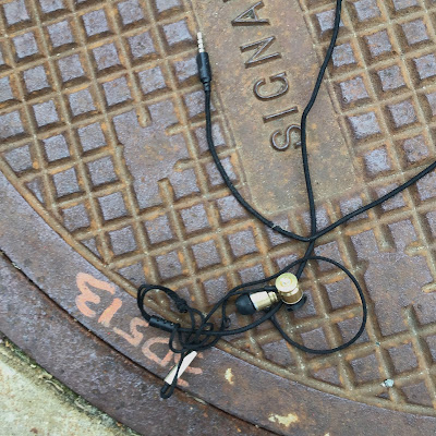 plug-in earbuds of black silicon earpiece and brass colored, bullet case-shaped body lying on cast-iron manhole cover