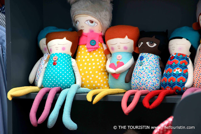 Eight colourful handmade dolls sitting on a wooden shelf, their spindly legs dangling in the air.