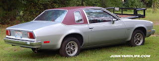 A silver 1975 Chevy Monza Towne parked on grass showing side and notchback trunk .