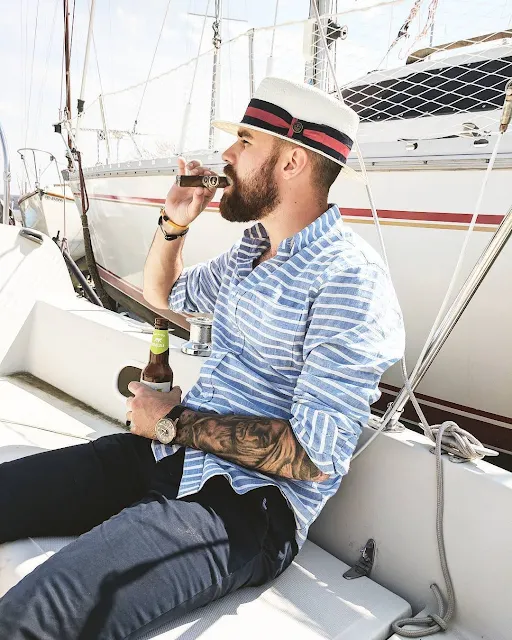 Bearded tattooed man with a white pork pie hat side view smoking a cigar on a boat
