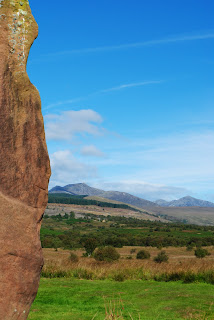 A photo showing on one side part of the red sandstone of a standing stone and then a view over the moor to the mountains in the background.  Photograph by Kevin Nosferatu for the Skulferatu Project.