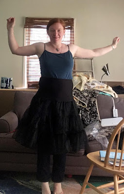 A young reheaded white woman, standing on tiptoe in a cluttered living room in front of a grey couch. Her arms are outstretched for balance and she's wearing a teal camisole, jeans, and a tea length black petticoat.