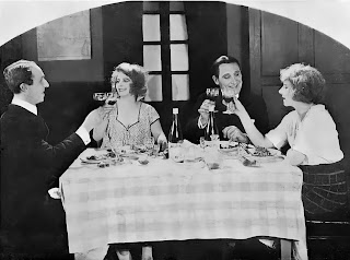 Deux hommes et deux femmes assis autour d'une table avec nappe à carreaux lèvent leur verre.
