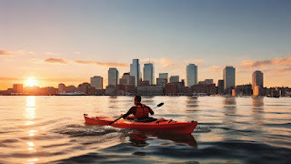 A person kayaking in Boston Harbor at sunset, with the city skyline glowing in the distance. The kayaker wears a life jacket and paddles through the calm water.