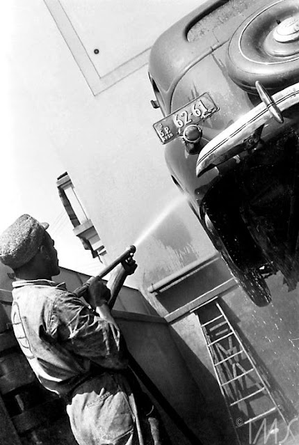 A fotografia, em preto e branco, retrata um homem lavando um carro. Ele utiliza uma boina, uma blusa de manga longa e calça de uniforme. Há um carro em suspensão, que é lavado por ele na altura da roda, por uma mangueira.