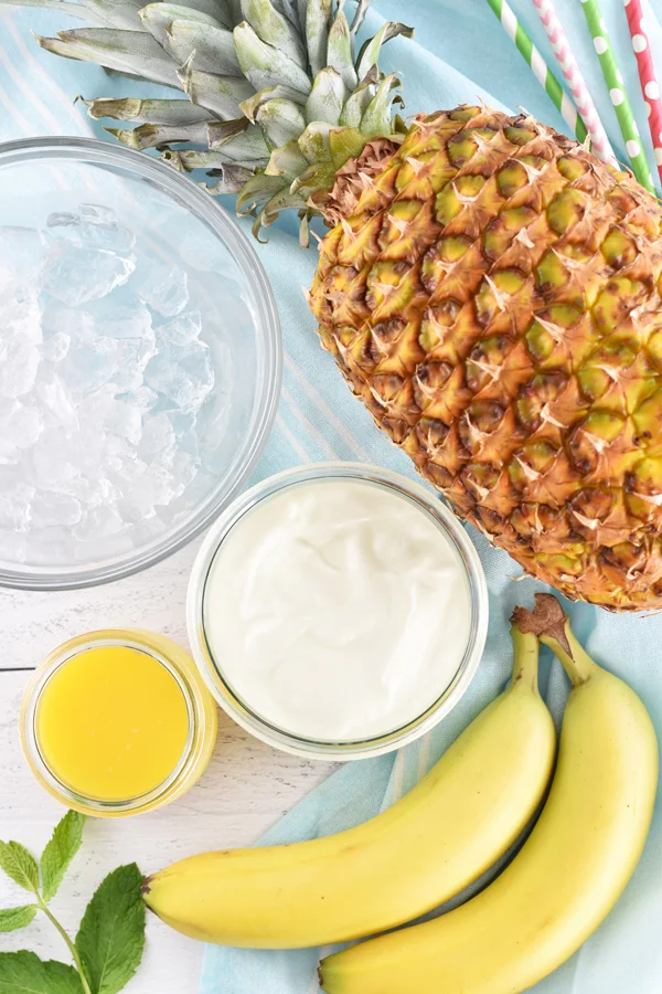op view photo of ingredients for pineapple banana smoothie, including ice, orange juice, pineapple, ripe bananas, yogurt, and fresh mint leaves.